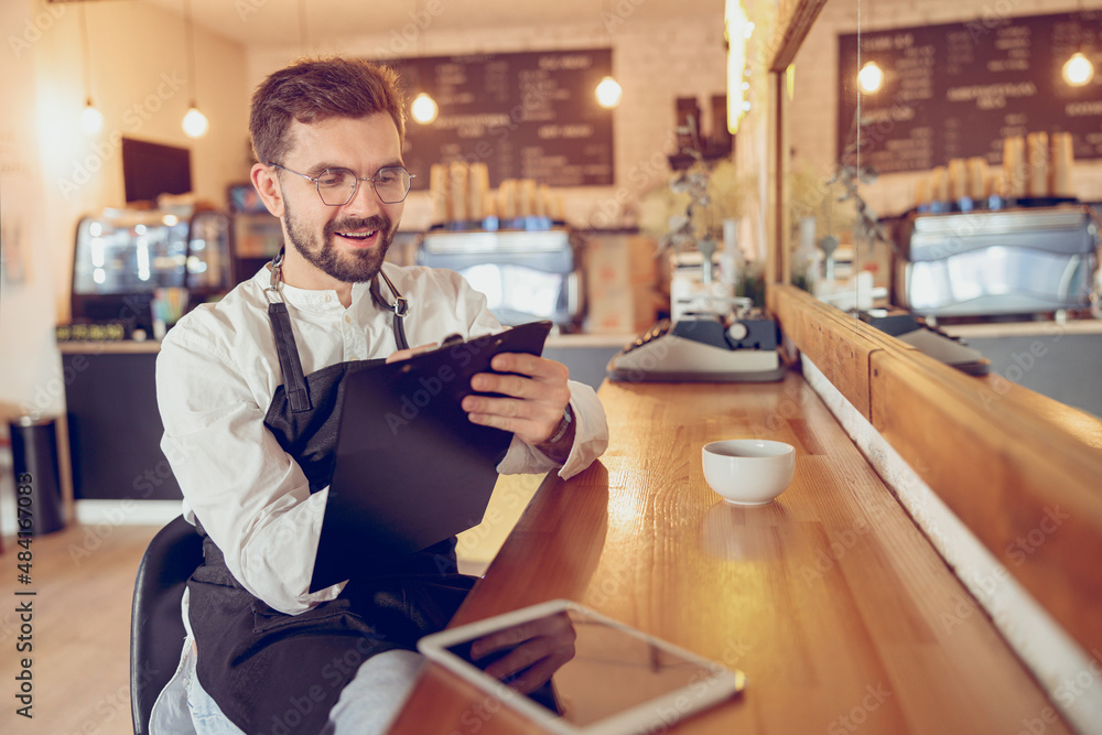 Fototapeta premium Joyful male barista writing on clipboard in cafe