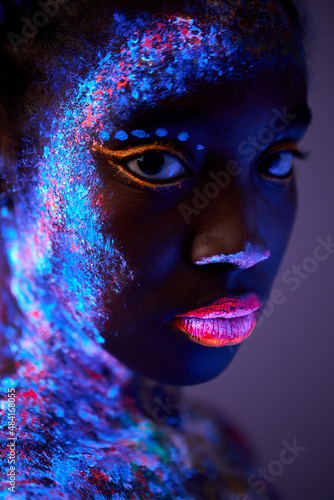 sensual photo of young stunning black woman with flurescent make-up on body posing in studio on ultraviolet light, neon. fashionable shoot of peaceful lady looking at camera photo