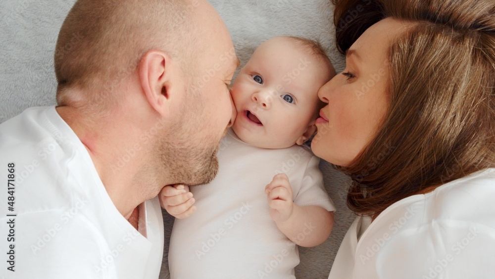 Happy family. Newborn baby with happy parents, top view. Healthy newborn baby in a white t-shirt with mom and dad. Close up Faces of the mother, father and infant baby. Cute Infant boy and parents Happy family. Newborn baby with happy parents, top view. Healthy newborn baby in a white t-shirt with mom and dad. Close up Faces of the mother, father and infant baby. Cute Infant boy and parents