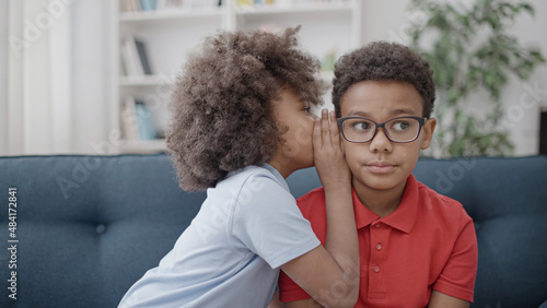 Little black girl whispering secret to brother in eyeglasses, siblings gossiping