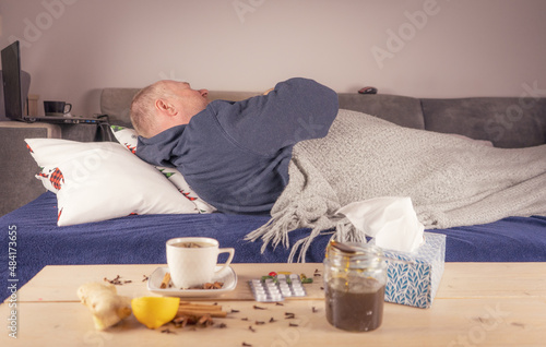 Mężczyzna z gorączką leży w łóżku na stole tabletki, miód, herbata, chusteczki, cytryna pomocne w leczeniu.