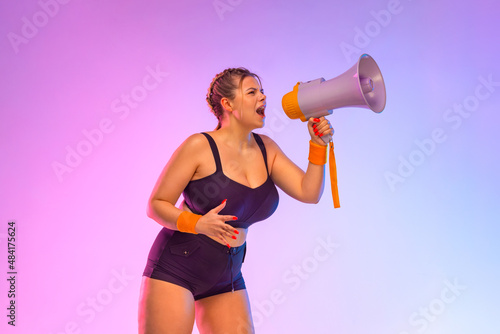 Emotional plus size woman in fitness sportswear screaming using megaphone. Social issues, protest concept. Side view. A girl speaks into a megaphone about an upcoming promotion and discounts in store © Mike Orlov