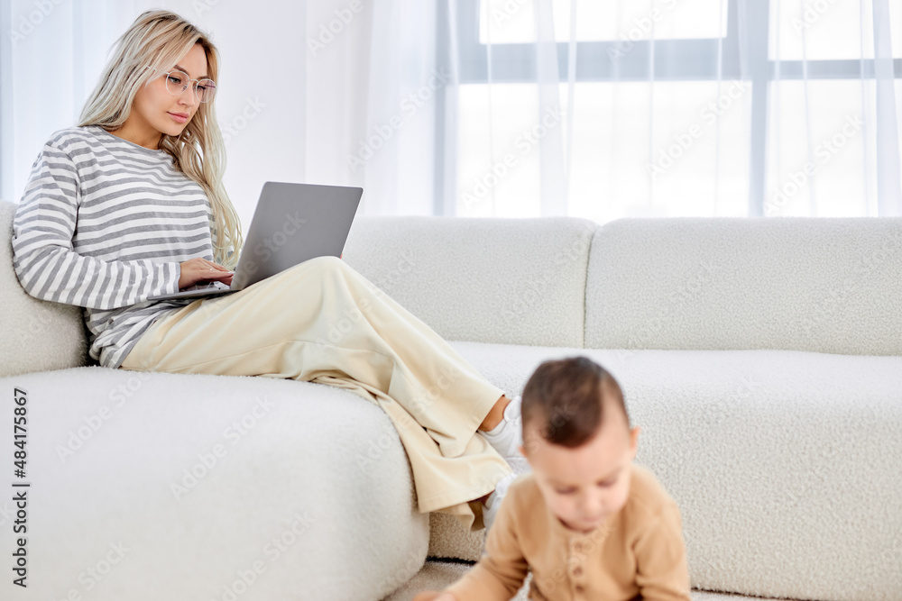 blonde caucasian mother working at home on sofa writing an email while child playing on floor. Young pretty woman working from home, while in quarantine isolation during the Covid-19 health crisis blonde caucasian mother working at home on sofa writing an email while child playing on floor. Young pretty woman working from home, while in quarantine isolation during the Covid-19 health crisis