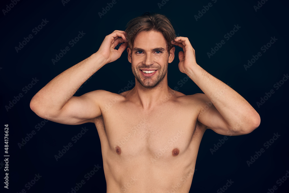 Handsome young man isolated. Portrait of shirtless muscular man is standing on dark blue background and holding his hair.