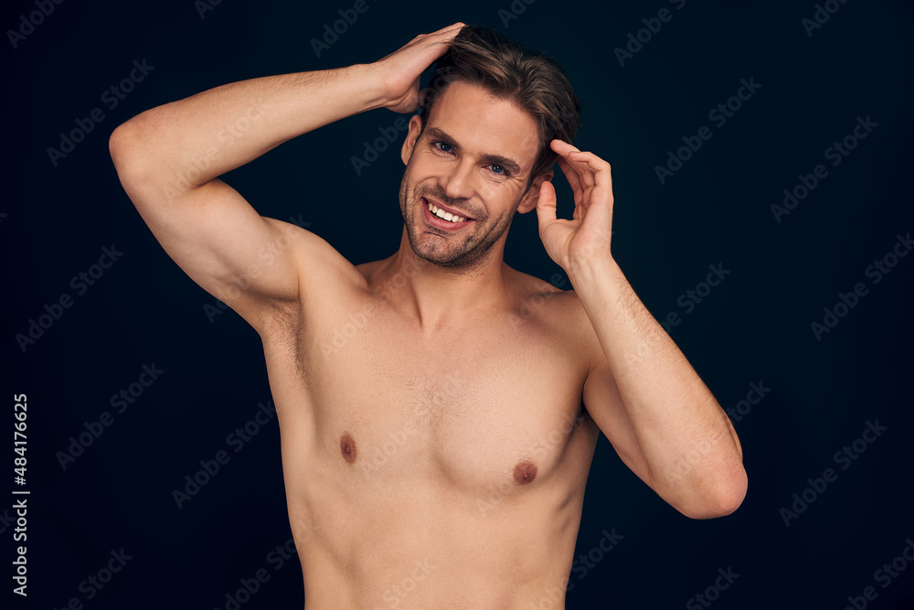 Fototapeta premium Handsome young man isolated. Portrait of shirtless muscular man is standing on dark blue background and holding his hair.