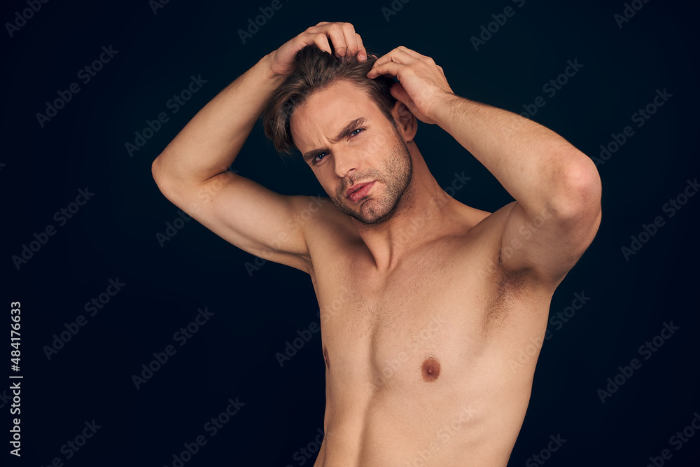 Handsome young man isolated. Portrait of shirtless muscular man is standing on dark blue background and holding his hair.