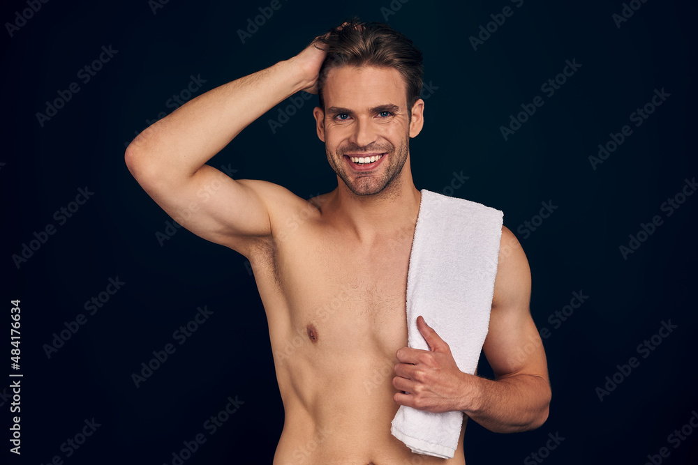 Fototapeta premium Handsome young man isolated. Portrait of shirtless muscular man is standing on dark blue background with towel and holding his hair.