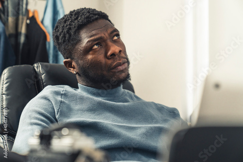 Thinking black man on black chair working on laptop looking up - portrait shot. High quality photo