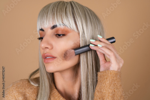 Blonde girl with bangs and brown daytime make-up holds a brush for blush contouring and highlighter photo