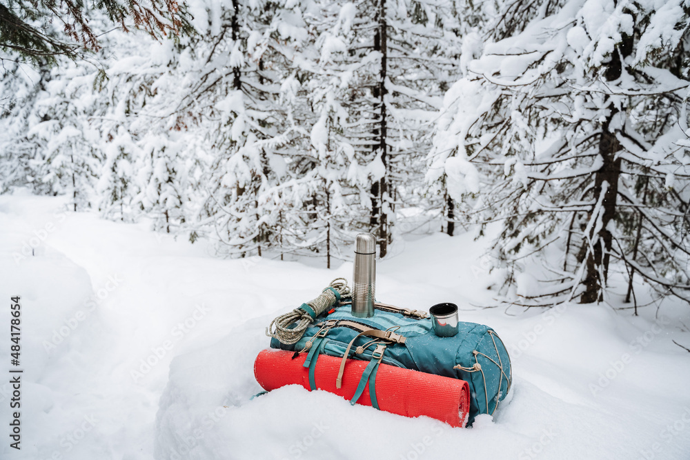 View of a backpack lying in the snow near the trail. The red mat is fastened with straps to the backpack. Thermos of silver color. A harsh winter hike through the forest.