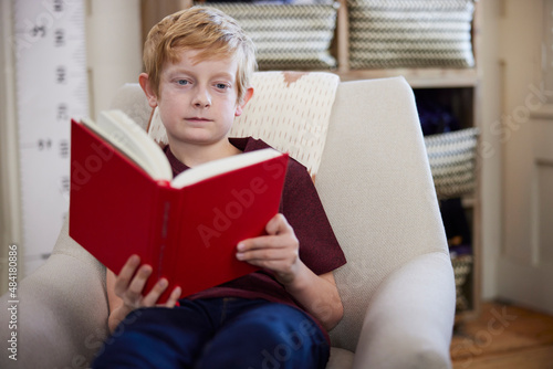 Boy Sitting In Chair In Bedroom At Home Reading Book