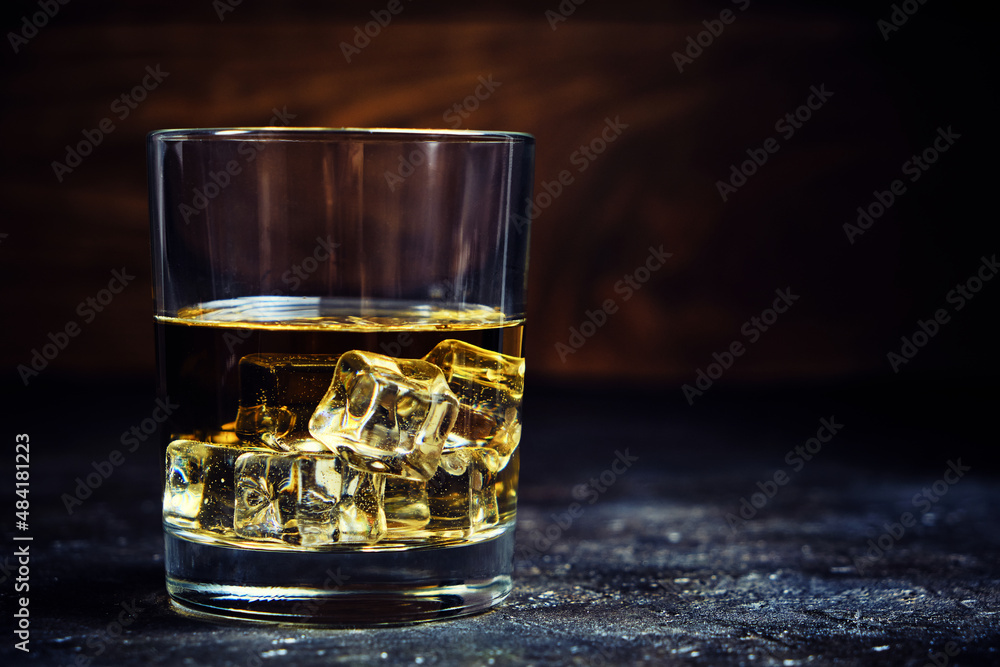 Glass chilled whiskey with ice cubes on wooden background in cellar.