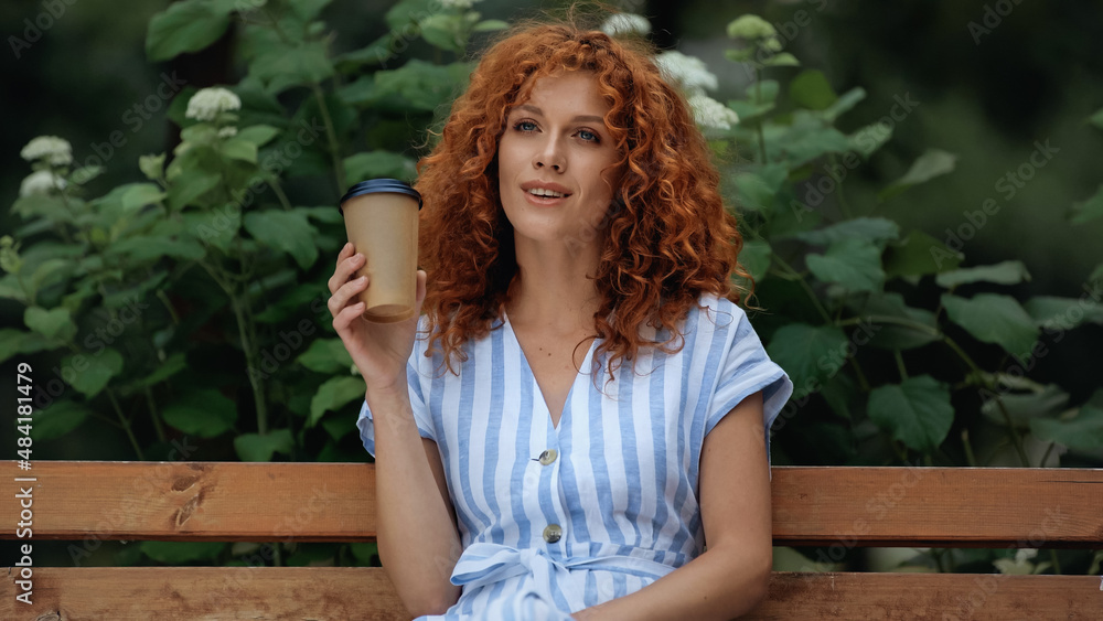 Naklejka premium pleased redhead woman in dress holding paper cup while sitting on bench in park.