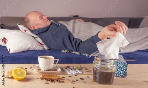 Mężczyzna z gorączką leży w łóżku. Na stole tabletki, herbata, chusteczki na katar, cytryna i miód. photo
