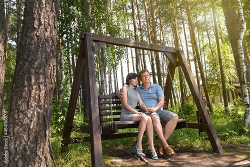An adult couple in love, a man and a woman, are sitting on a wooden swing in the forest enjoying nature on a summer day.