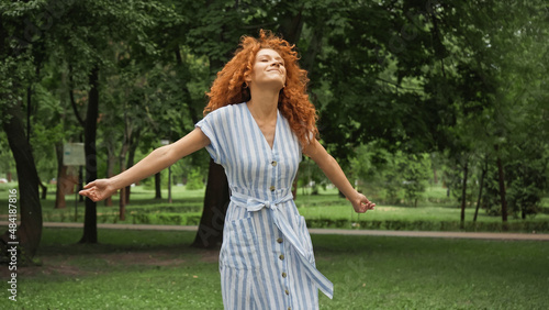 pleased young woman with red hair standing with outstretched hands in green park.