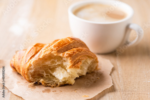 Half eaten fresh french croissant and cup of coffee on background, wooden table background. Coffee break in cafe