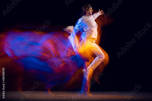 Youth dances. Dynamic portrait of hip-hop dancer in action, motion isolated on dark background in mixed neon light. Youth culture, hip-hop, style and fashion