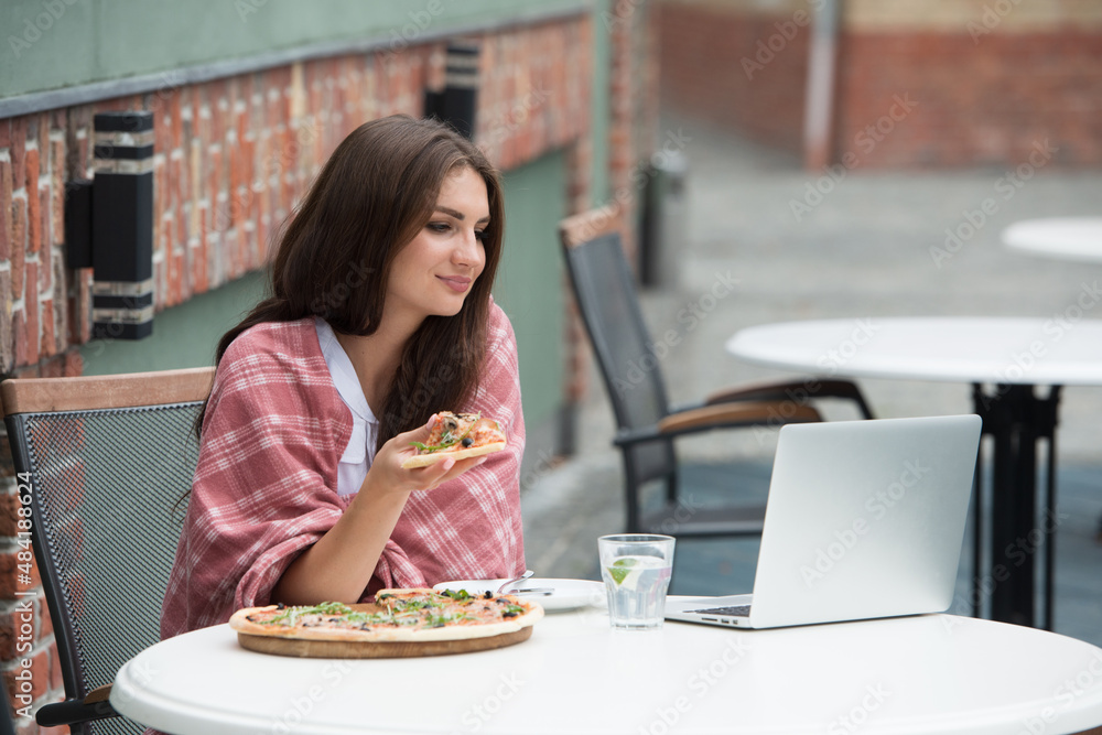 Young freelancer woman using laptop computer and eat pizza while sitting at cafe table.