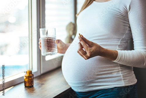 Pregnant woman reading label on bottle with medicine, with vitamins. Female sitting at home in bed with glass of water medicine. Pregnancy, health, pharmaceuticals, care and people.