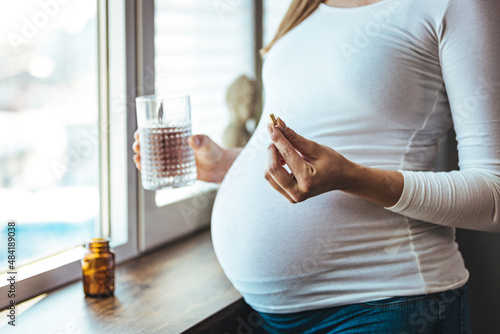 Pregnant woman reading label on bottle with medicine, with vitamins. Female sitting at home in bed with glass of water medicine. Pregnancy, health, pharmaceuticals, care and people. photo