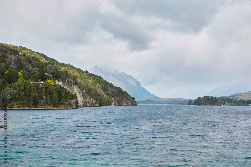 Lake in Argentinian Patagonia with turquoise water and trees in the background between mountains. Cloudy day with clouds between the mountains.