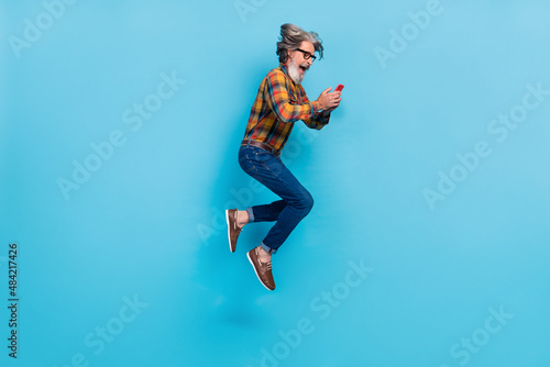 Photo of excited retired man wear plaid shirt spectacles jumping high typing modern device isolated blue color background