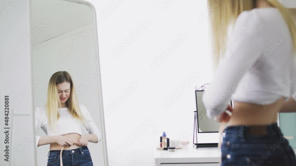A young woman stands in front of a mirror and measures the circumference of her waist with a tape measure. The girl is complexed because of the weight A young woman stands in front of a mirror and measures the circumference of her waist with a tape measure. The girl is complexed because of the weight