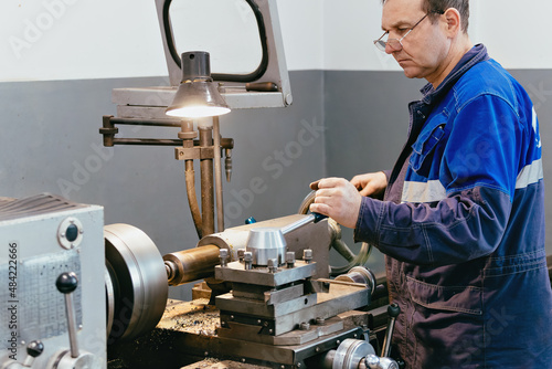 Adult turner of 50-55 years old works in workshop behind lathe. Industrial production of metal parts. Caucasian worker in overalls. Authentic work process scene in production. Real worker. Not staged photo