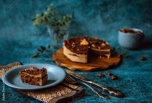 Homemade cake with chocolate and cream on round wooden board, plate with piece of cake next to fork and knife and mug filled with nuts on dark background. Home baking