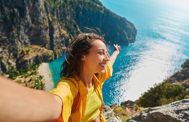 Positive tourist woman taking picture outdoors for memories, making selfie on top of cliff with valley mountains view, sharing travel adventure journey