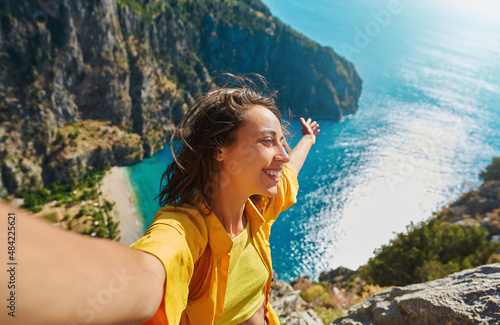 Positive tourist woman taking picture outdoors for memories, making selfie on top of cliff with valley mountains view, sharing travel adventure journey