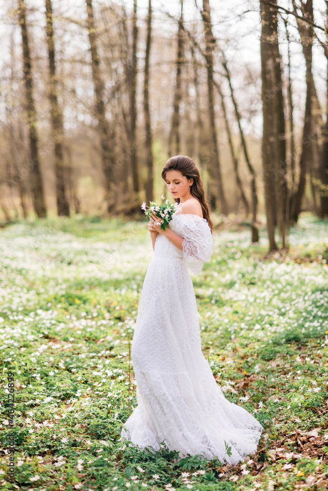 young beautiful woman in a white long dress collects primroses. Fairy girl in the spring forest. A bouquet of white flowers in her hands