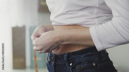 A young woman stands in front of a mirror and measures the circumference of her waist with a tape measure. Close-up photo