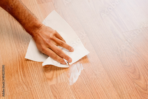 A man's hand wiping a white cream stain on the wooden floor with a tissue.