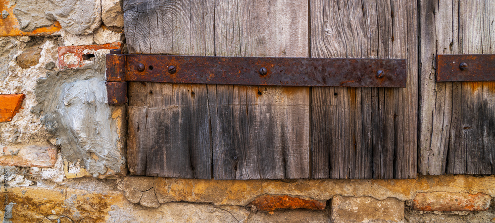 Part weathered wooden door with steel hinges. Distressed wood panels. Castle stone wall texture background. Detail of an old wooden door with paint residues and rusty door latch. Part weathered wooden door with steel hinges. Distressed wood panels. Castle stone wall texture background. Detail of an old wooden door with paint residues and rusty door latch.