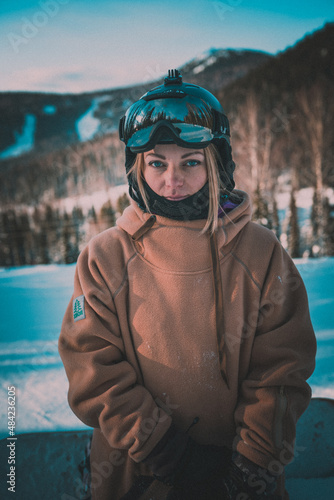 Portrait Beautiful young girl at a ski resort against the backdrop of snowy mountains. She is dressed in winter clothes for snowboarding and ski goggles. Winter travel at the ski resort.