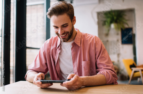 Cheerful hipster guy enjoying time for playing virtual 3d via nintendo console joystick while waiting friend, smiling male gamer passing next level via handheld joypad app during weekend leisure