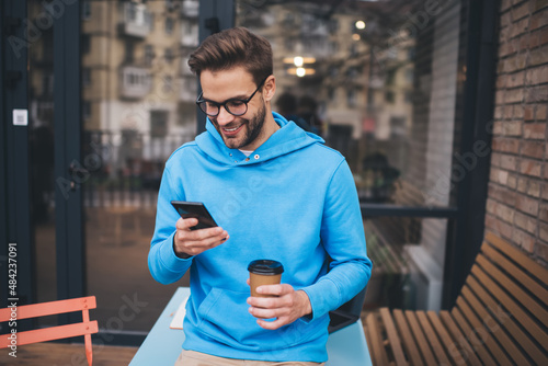 Cheerful hipster guy with takeaway coffee to go enjoying mobile messaging during coffee time on weekend, happy Caucaisan man in classic eyeglasses connecting to 4g wireless for making online booking © BullRun