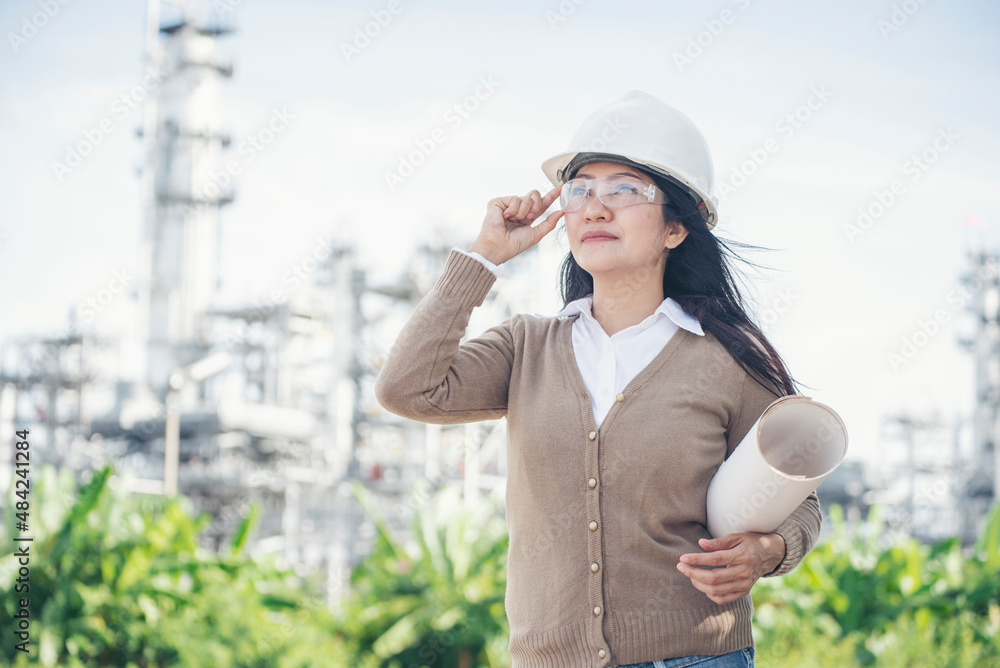 Young woman engineer or construction worker wearing safety hat (helmet) and holding blueprint with refinery plant background.
