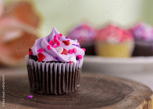 A chocolate valentine's day cupcake on a wooden slab and tulips and additional cupcakes blurred in the background. Light green background.