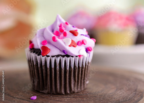 A chocolate valentine's day cupcake on a wooden slab and tulips and additional cupcakes blurred in the background. Light green background.