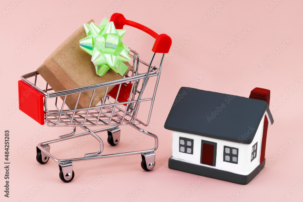Shopping cart and house on beige background. Real estate sale or purchase concept Shopping cart and house on beige background. Real estate sale or purchase concept