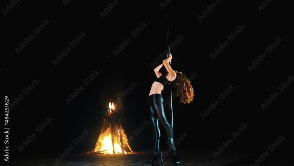 pole dance. witch halloween. fit female gymnast, in black leather outfit and high heels, performs acrobatic exercises on metal rotating pole, at night, by light of large fire in background. pole dance. witch halloween. fit female gymnast, in black leather outfit and high heels, performs acrobatic exercises on metal rotating pole, at night, by light of large fire in background.
