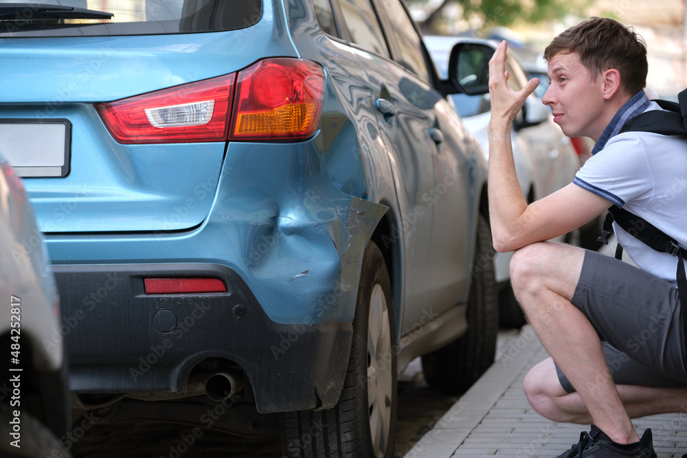Sad male driver sitting on street side shocked looking at his dented car parked at street side. Road safety and vehicle insurance concept Sad male driver sitting on street side shocked looking at his dented car parked at street side. Road safety and vehicle insurance concept