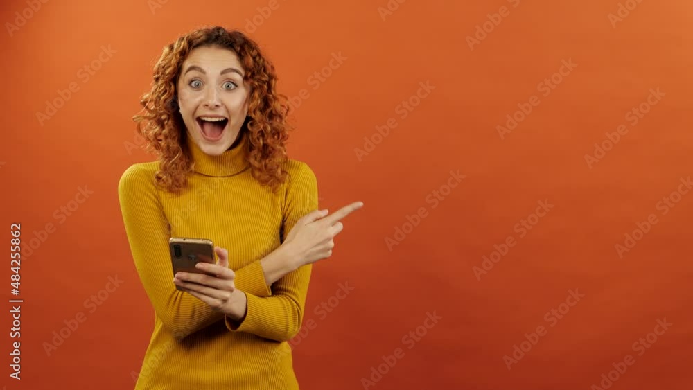 Attractive redhead caucasian girl in orange jumper is using mobile phone and shocked pointing finger at copy space for advertising isolated on orange background. Attractive redhead caucasian girl in orange jumper is using mobile phone and shocked pointing finger at copy space for advertising isolated on orange background.
