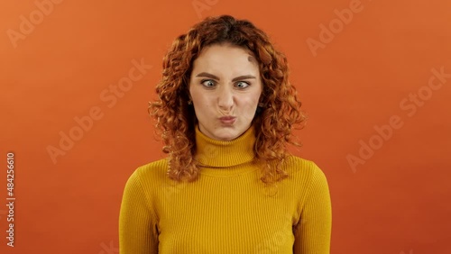 Attractive caucasian redhead girl in orange jumper making a stupid and comic face isolated on orange studio background.