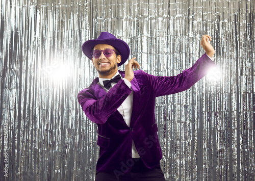 Happy goofy young man wearing a ridiculous tacky unfashionable purple velvet jacket and hat doing funny dance moves at a party. Hilarious dorky guy dancing to music on a silver foil fringe background photo