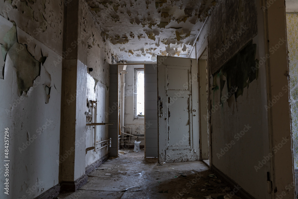 Obraz premium Dark corridor in an abandoned public building with an open creepy door. Urban exploration in an old horror house. Paint is peeling off the walls and dirt is lying on the floor. Old pipes are visible.