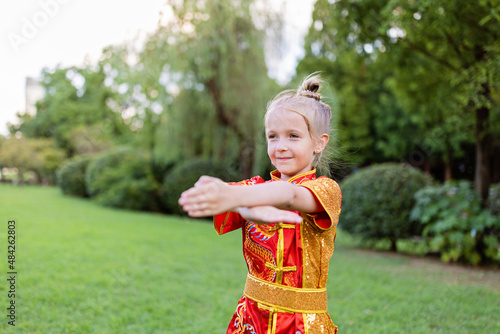 Cute little caucasian girl seven years old in red sport wushu uniform exercising in park at summer day. Lifestyle portrait of kung fu fighter child athlete photo